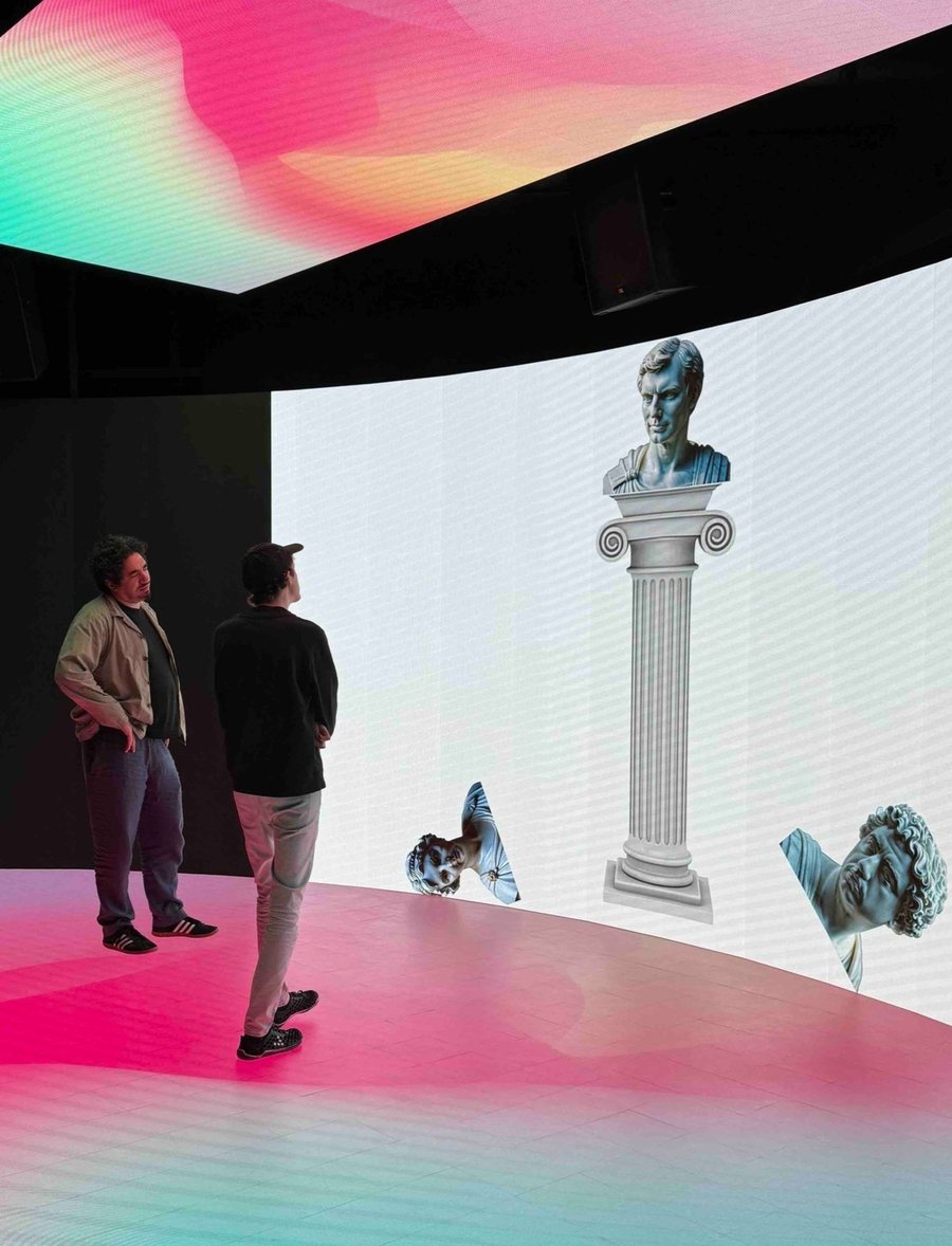 Two visitors in an immersive room with a classical bust on a column and digital sculptures on a curved screen
